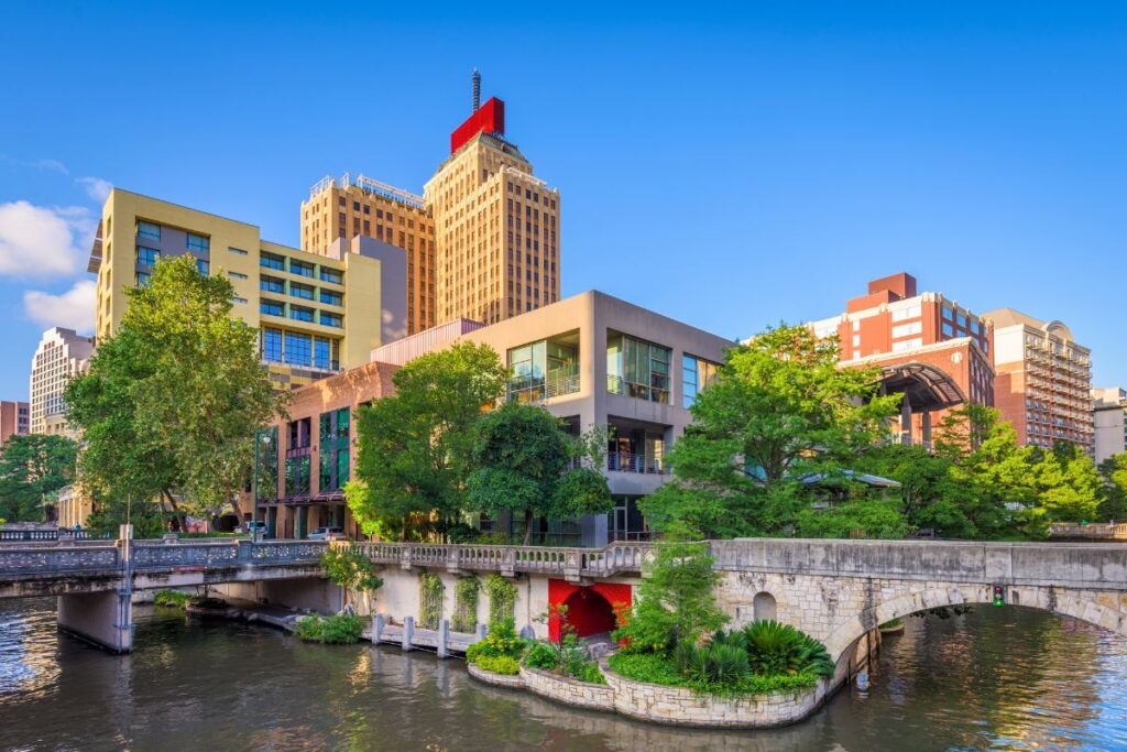 Scenic urban riverwalk featuring historic stone bridges crossing a peaceful waterway, surrounded by lush green trees, modern buildings, and high-rise architecture beneath a clear blue sky.