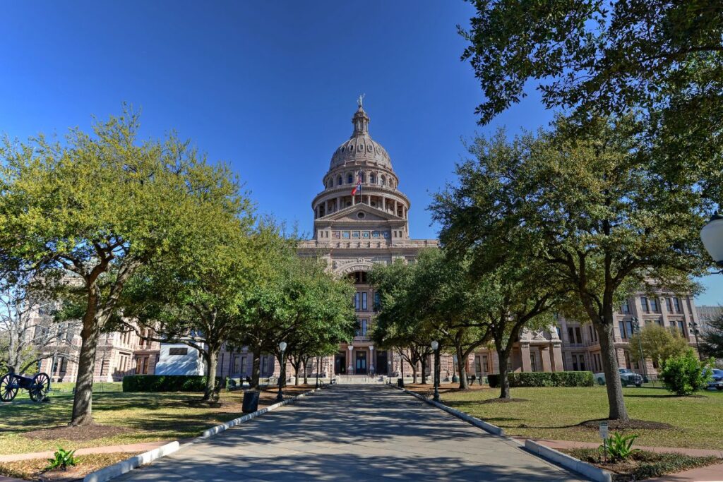 Historic limestone capitol building featuring an ornate domed roof, classical architecture, and a grand entrance framed by mature trees, landscaped lawns, and a paved walkway beneath a clear blue sky.