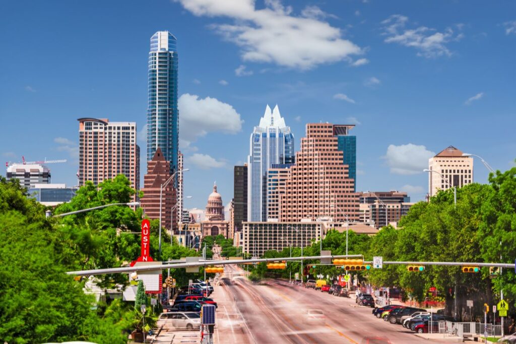 Wide downtown roadway lined with lush green trees, featuring modern skyscrapers, historic architecture, traffic lights, and a vibrant city skyline beneath a bright blue sky.